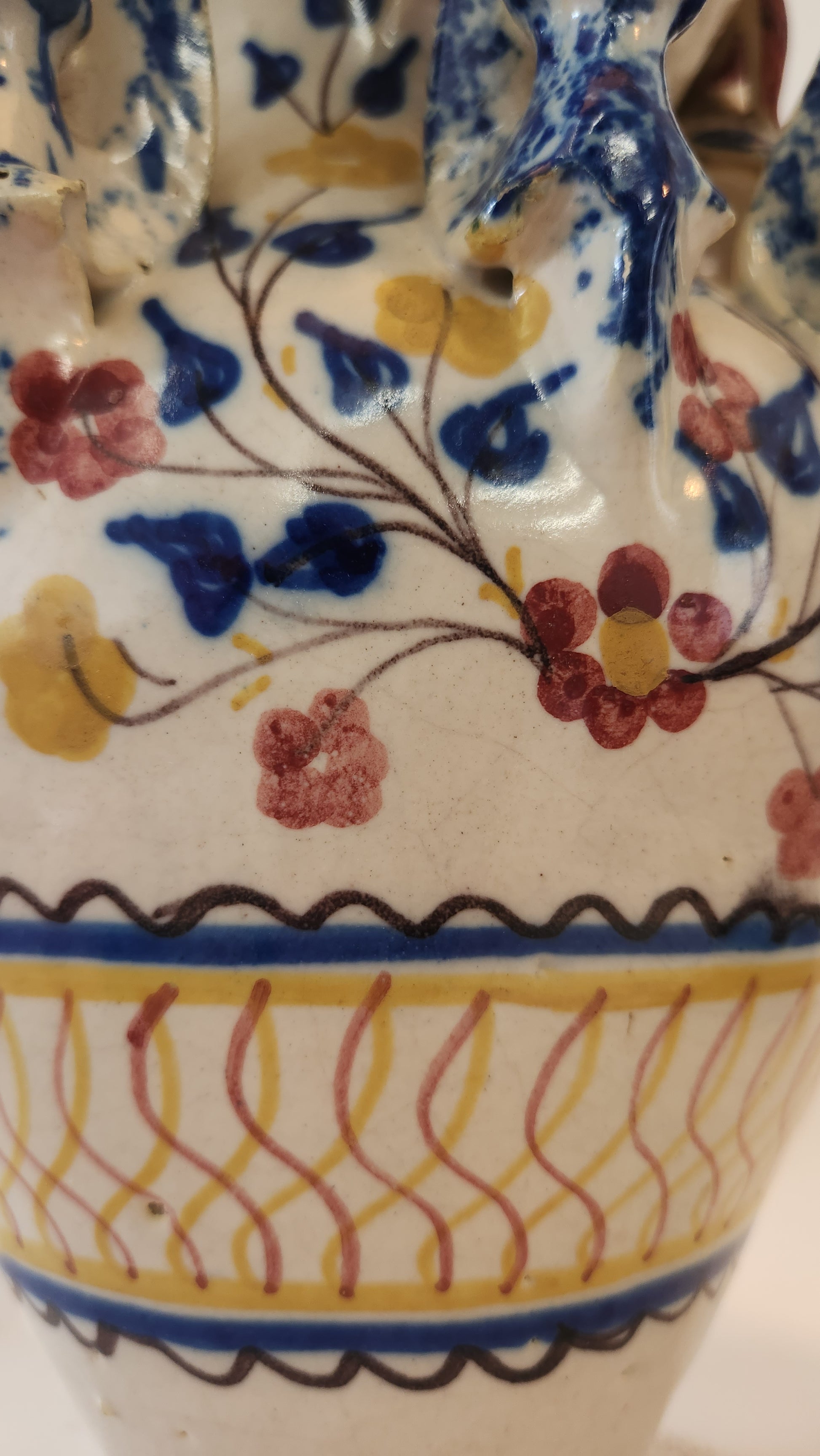 Close-up of a ceramic vase with a white glaze, decorated with blue floral motifs, red and yellow flowers, and a yellow wavy stripe bordered by black scalloped lines on the side.