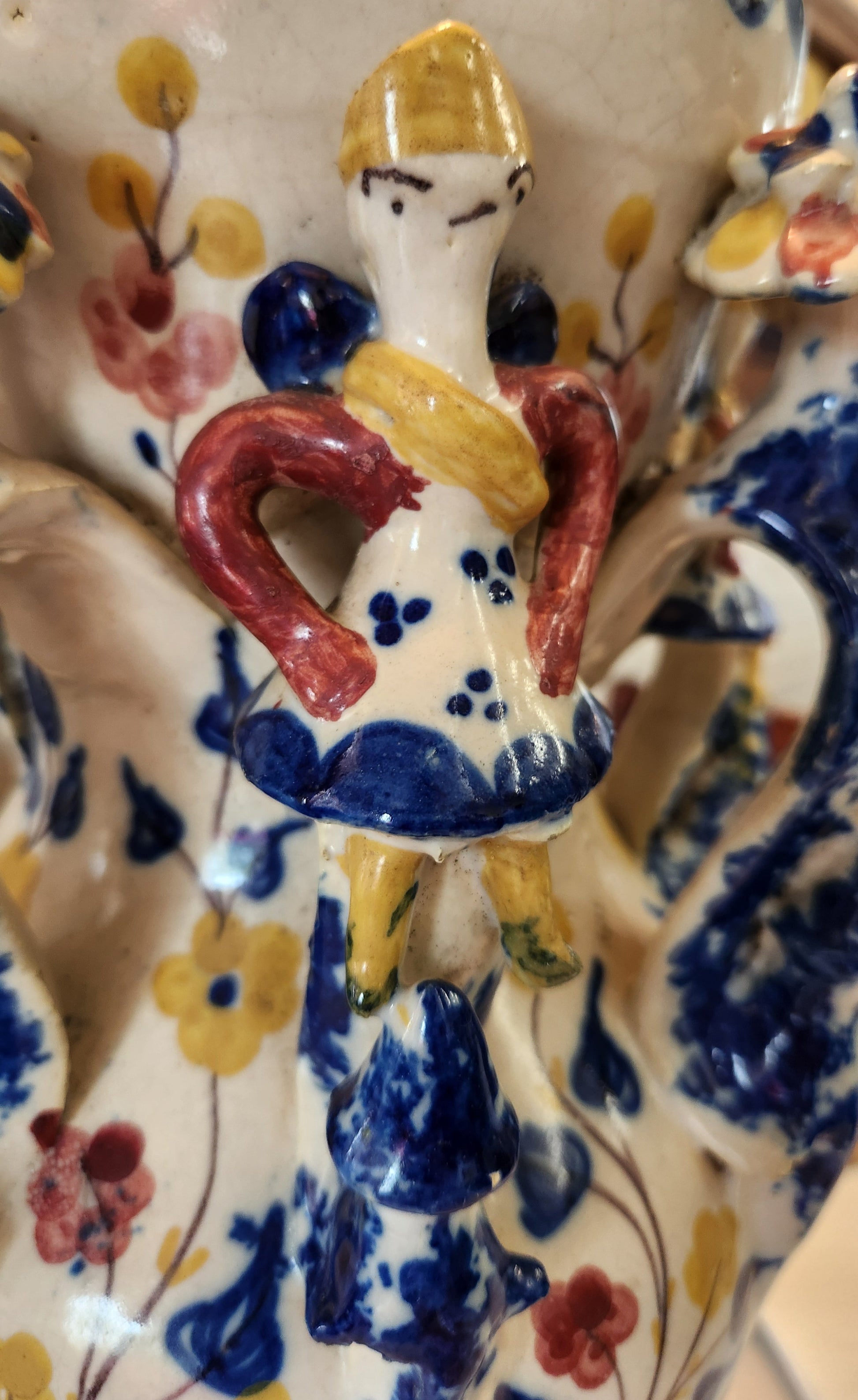 Close-up of a hand-painted ceramic plate or dish featuring a whimsical character with a yellow hat, blue body, and red arm, surrounded by floral patterns in blue, yellow, and red on an off-white background.