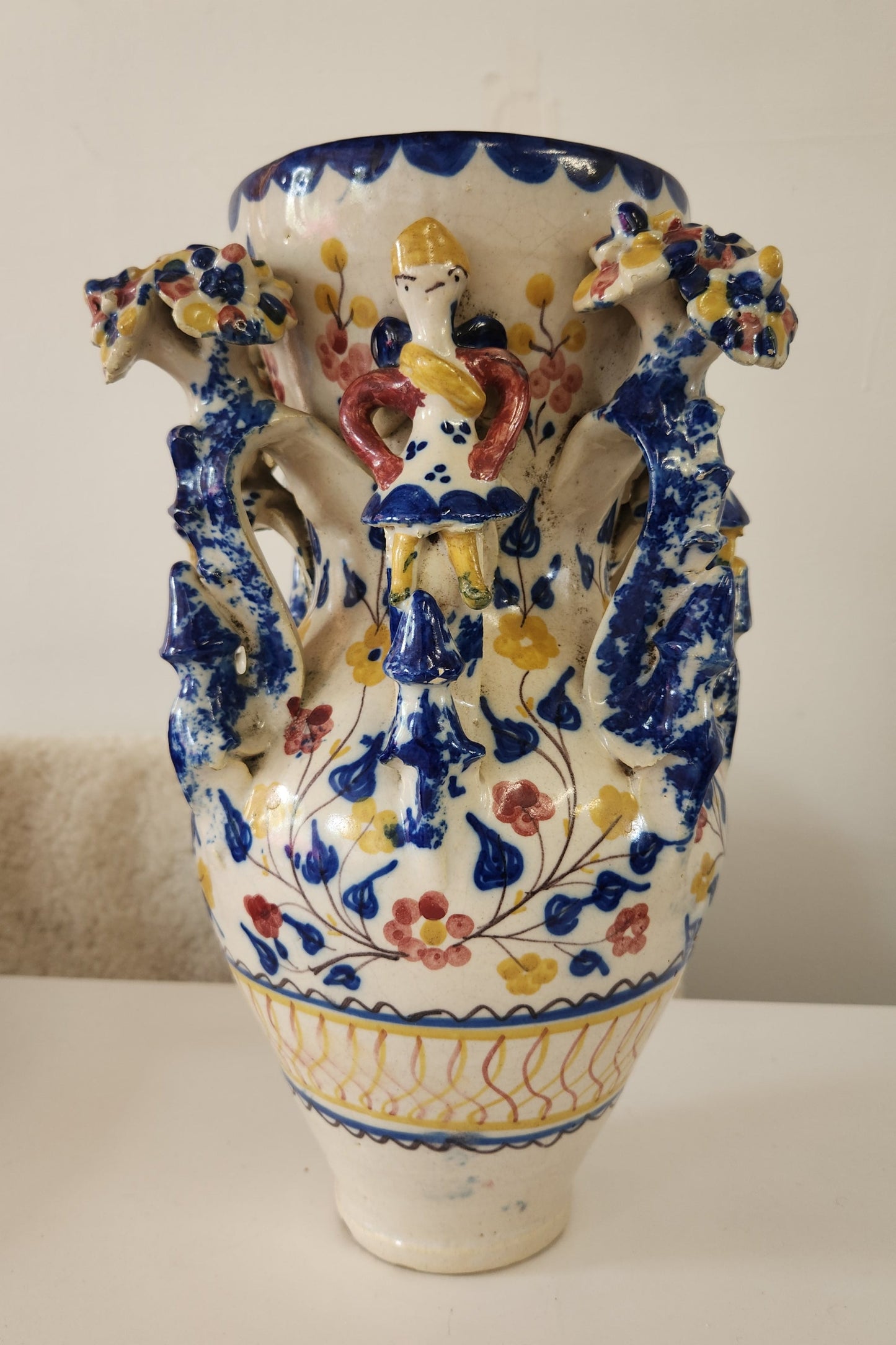 An ornate ceramic vase with a white base, decorated with blue floral patterns and raised multicolor figures in yellow, red, and green, photographed against a plain off-white background.