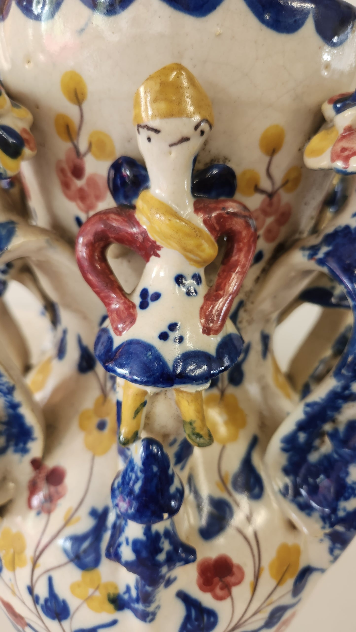 A close-up of a hand-painted ceramic plate featuring whimsical, stylized figures in yellow, red, and blue on an off-white background with floral patterns.