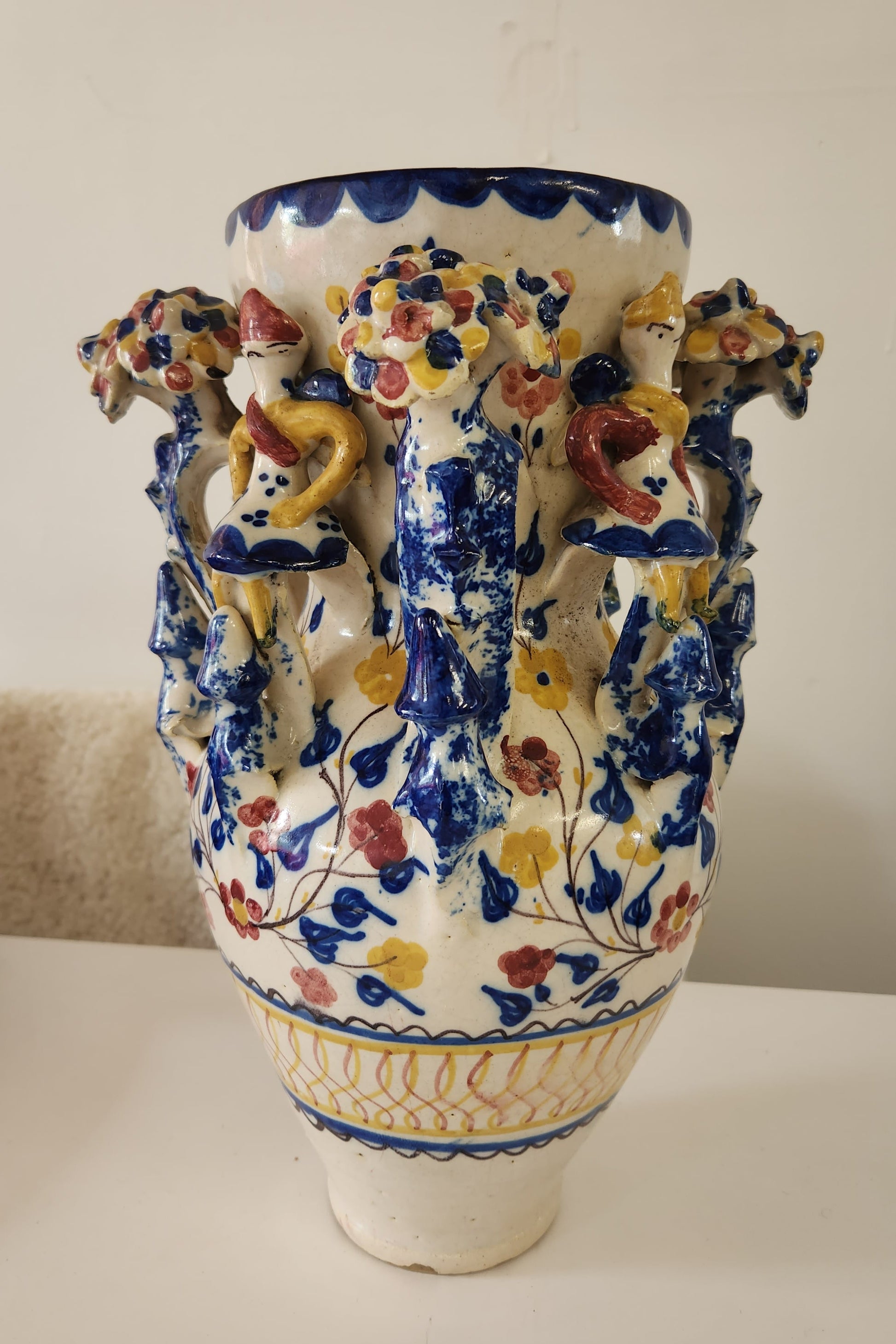 An ornate ceramic vase with a white base, decorated with blue floral patterns and raised multicolored animal figures, photographed against a plain light background.