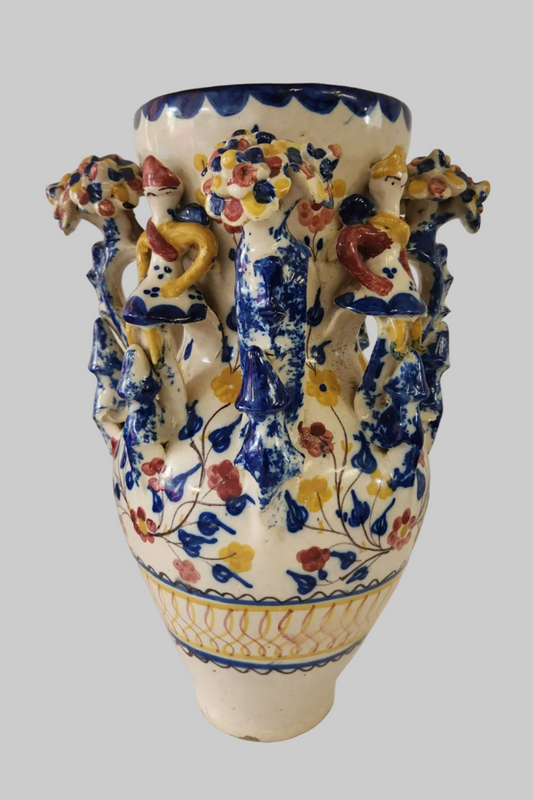 An ornate ceramic grotesque jar vase with a cream-colored glaze, decorated with blue floral patterns and raised multicolored figures around the sides, shown against a solid gray background.