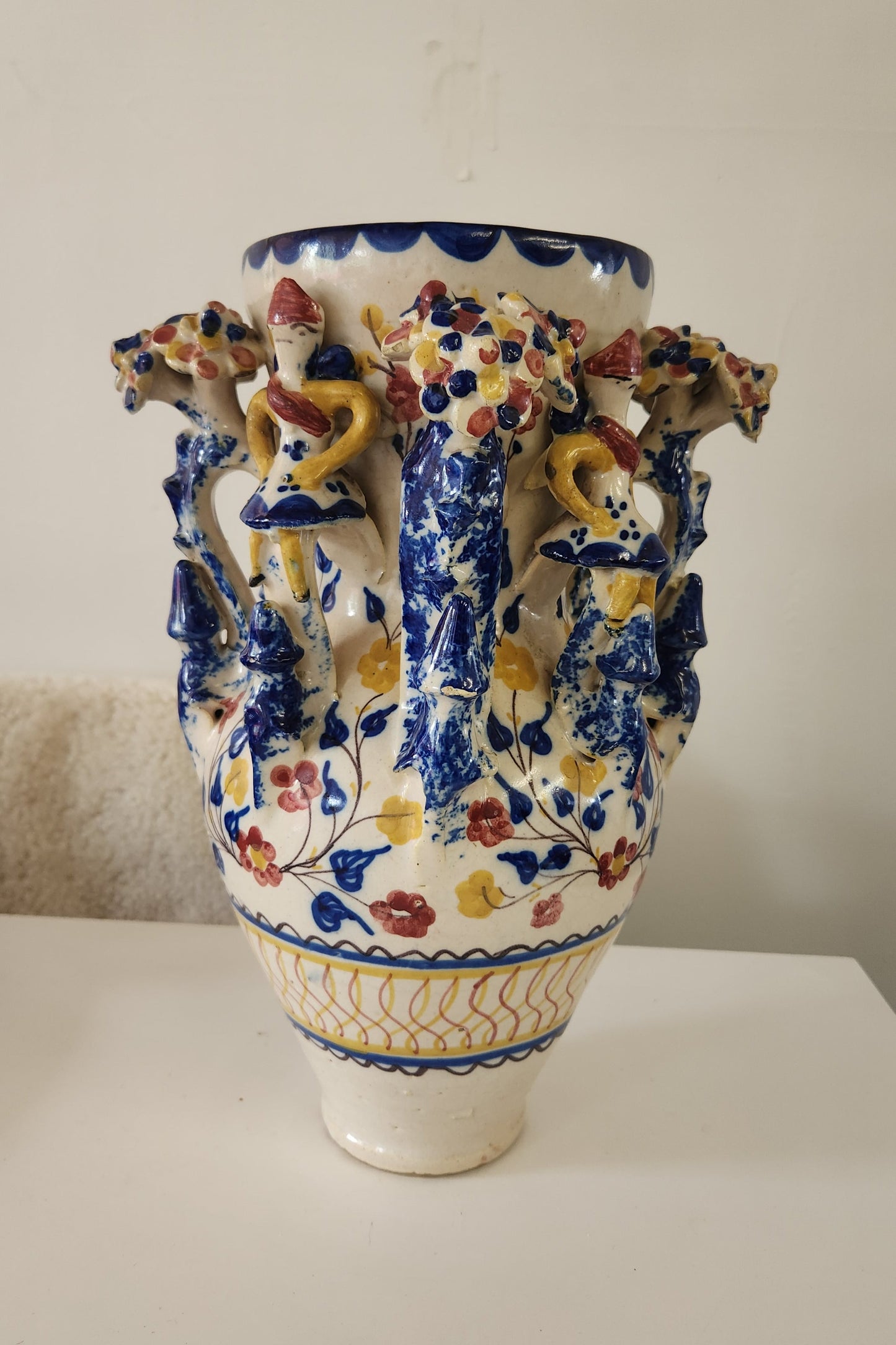 A decorative ceramic vase with a white base, featuring a blue floral pattern and three sculpted figures in yellow, red, and blue on the shoulder, photographed against a plain off-white wall.