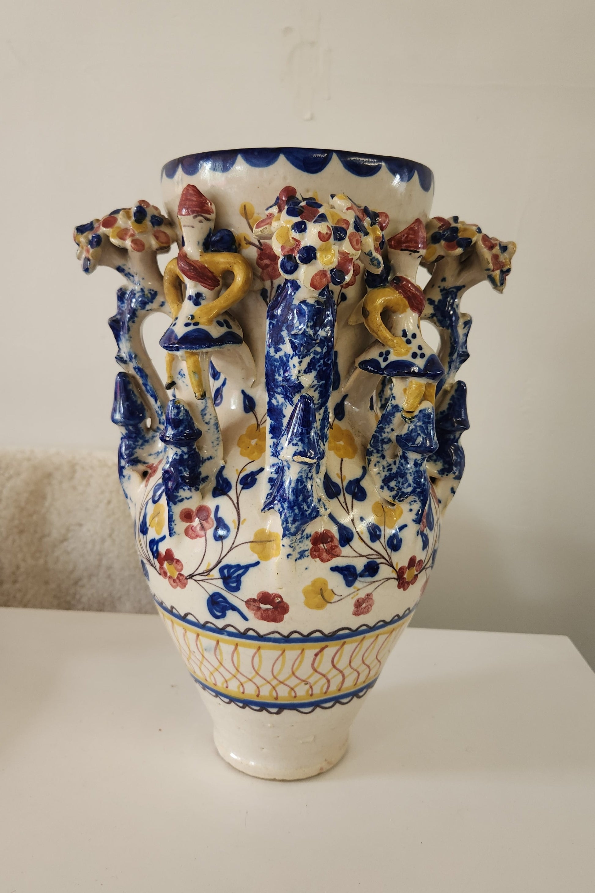 A decorative ceramic vase with a white base, featuring a blue floral pattern and three sculpted figures in yellow, red, and blue on the shoulder, photographed against a plain off-white wall.