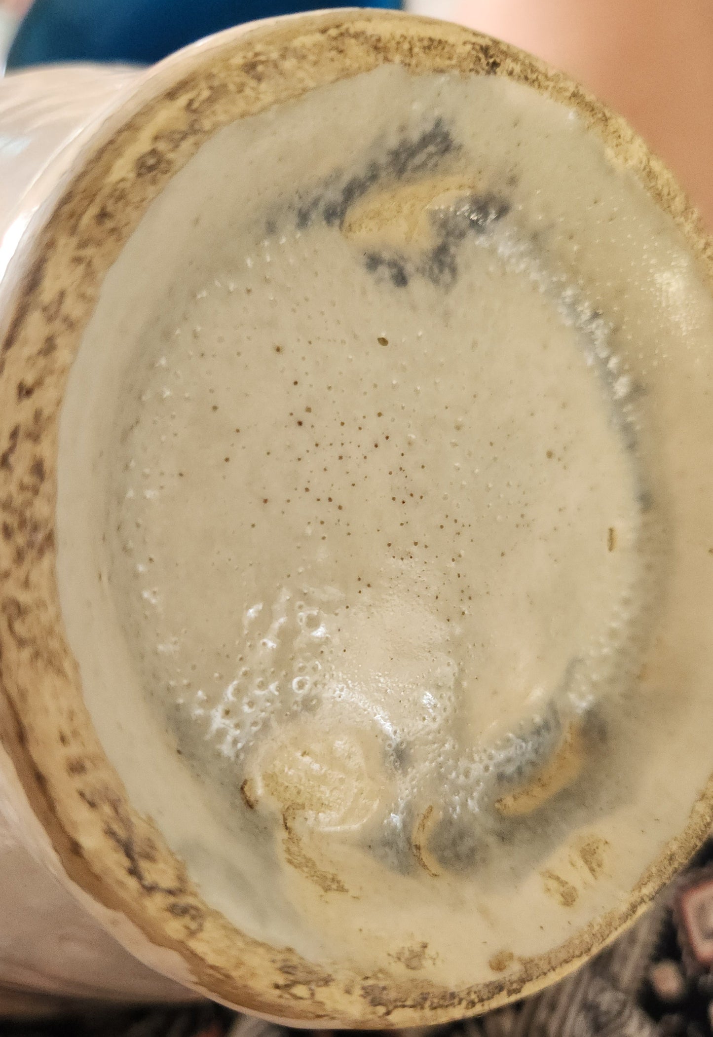 Close-up of the bottom interior of a ceramic cup or vase, showing a textured off-white glaze with dark speckles and a rustic brown rim.