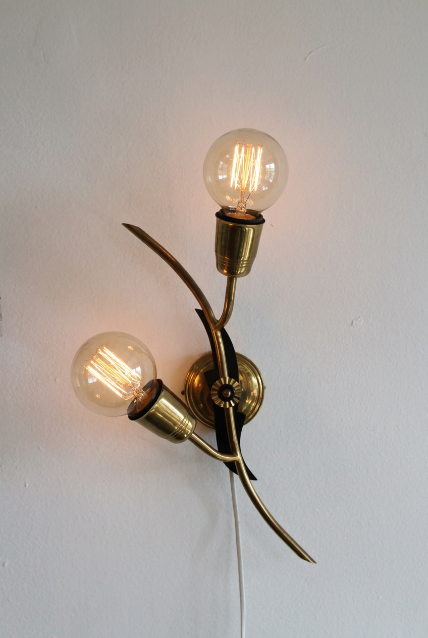 A modern wall-mounted light fixture with two curved brass arms, black accents, and two frosted glass bulbs against a plain white wall.