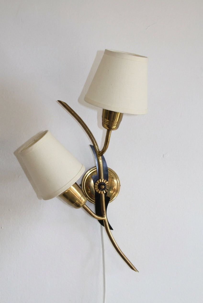 A vintage-style brass & black isolit wall sconce with a cream fabric lampshades, mounted on a plain white wall.