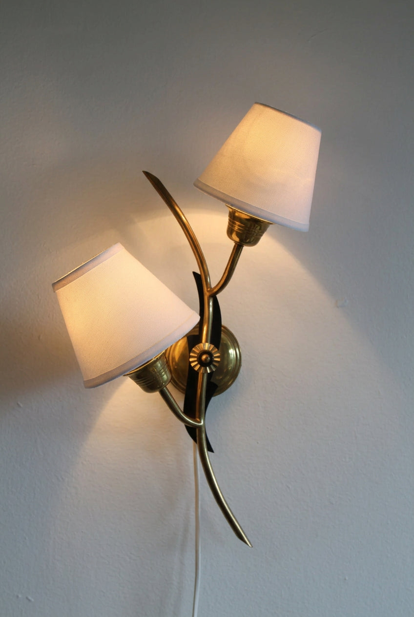 A two-light wall sconce with warm white fabric shades and a polished brass/gold metal frame, mounted on a plain light gray wall.