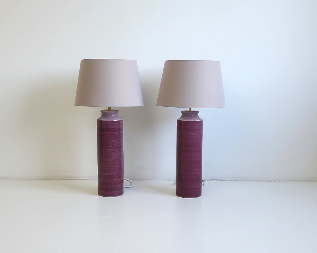 Two table lamps with purple cylindrical bases and beige lampshades on a concrete floor.