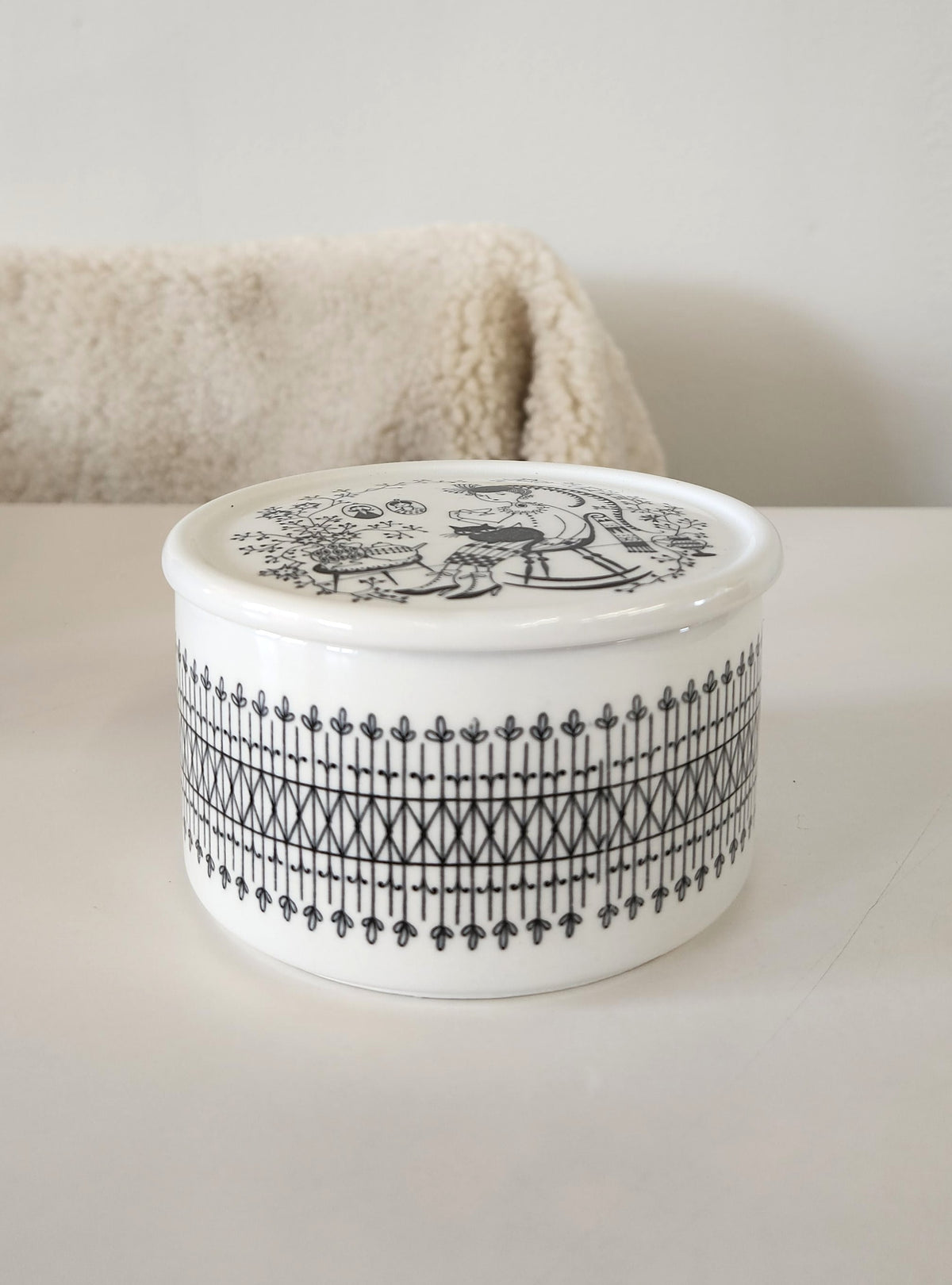 A round white ceramic lidded box made by Arabia, with black hand painted patterns by Raija Uosikkinen, including a decorative border and a portrait illustration on the lid of a woman with a fat cat on her lap, set against a neutral white background. 