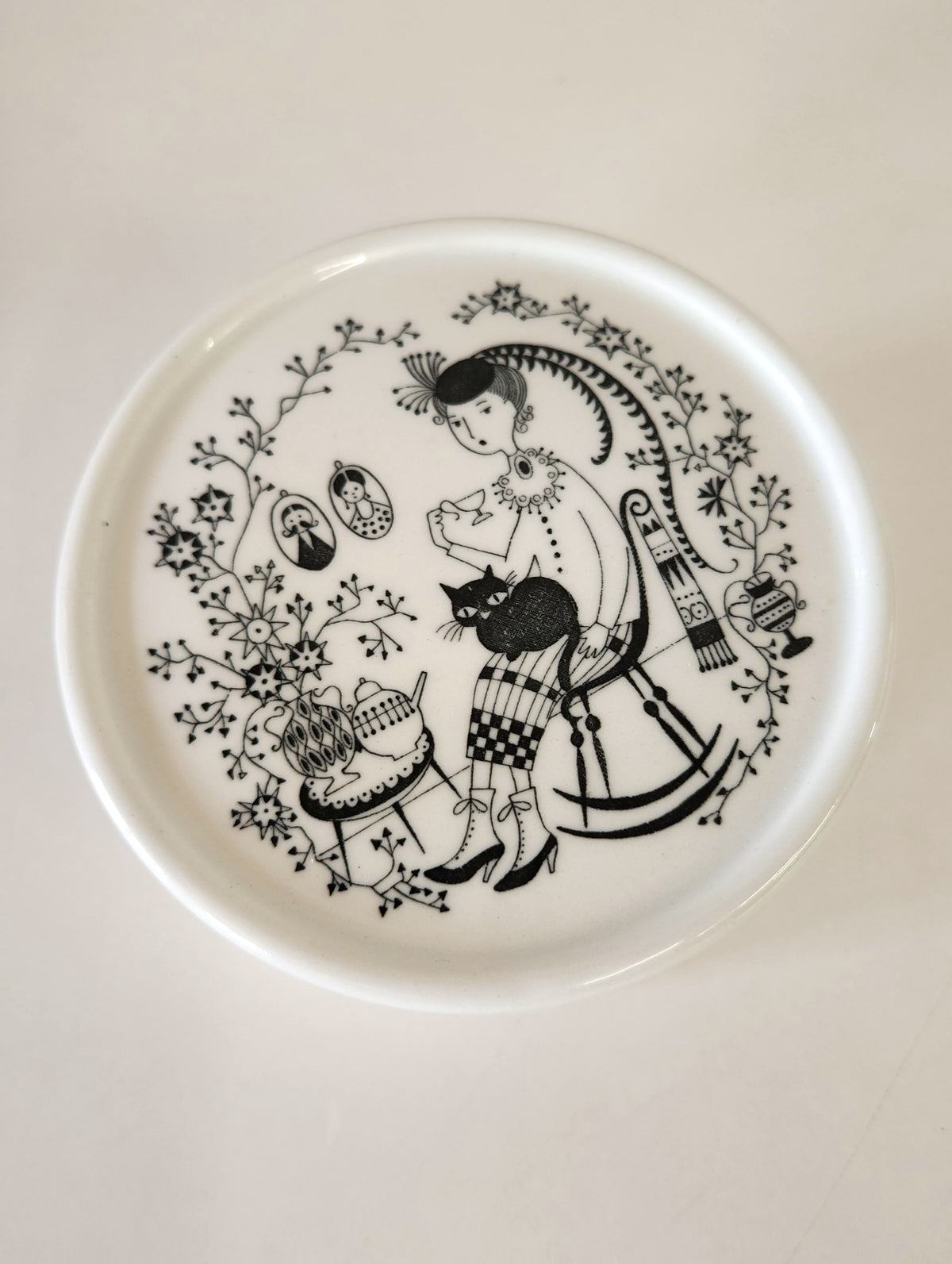A round white ceramic lidded box made by Arabia, with black hand painted patterns by Raija Uosikkinen, including a decorative border and a portrait illustration on the lid of a woman with a fat cat on her lap, set against a neutral gray background. 