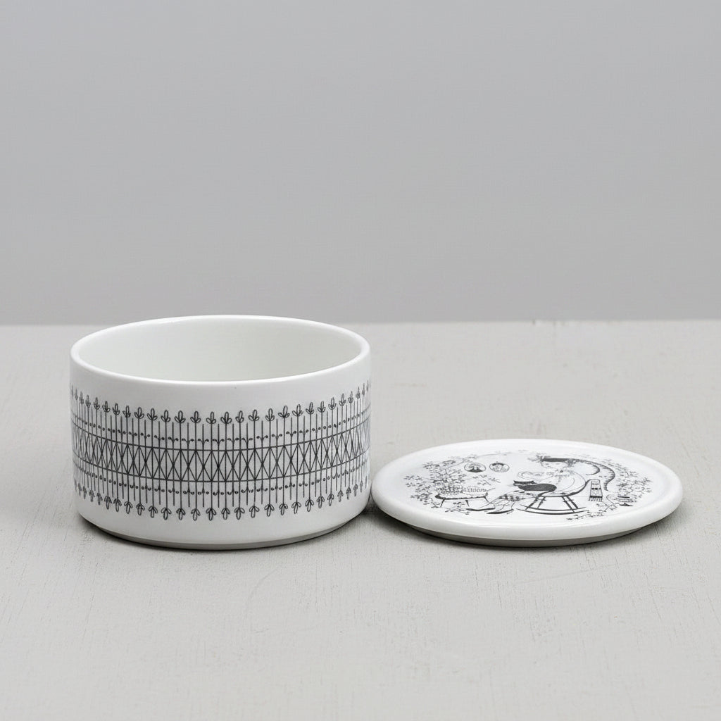 A round white ceramic lidded box made by Arabia, with the lid off. Black painted patterns, including a decorative border and a portrait illustration on the lid of a woman with a fat cat on her lap, set against a neutral gray background. 