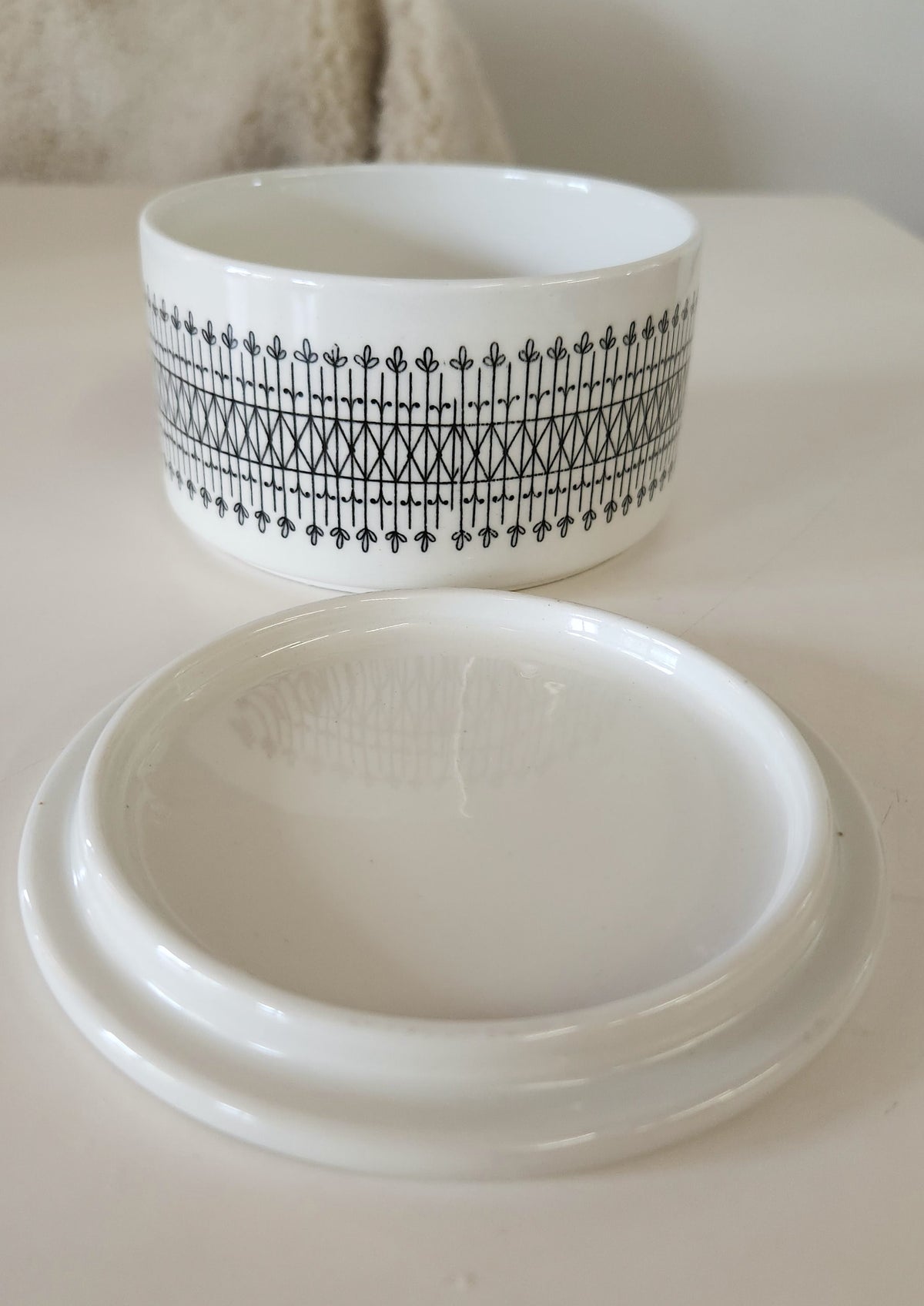 A round white ceramic lidded dish made by Arabia, with the lid off.  Jar has a black hand painted decorative border 