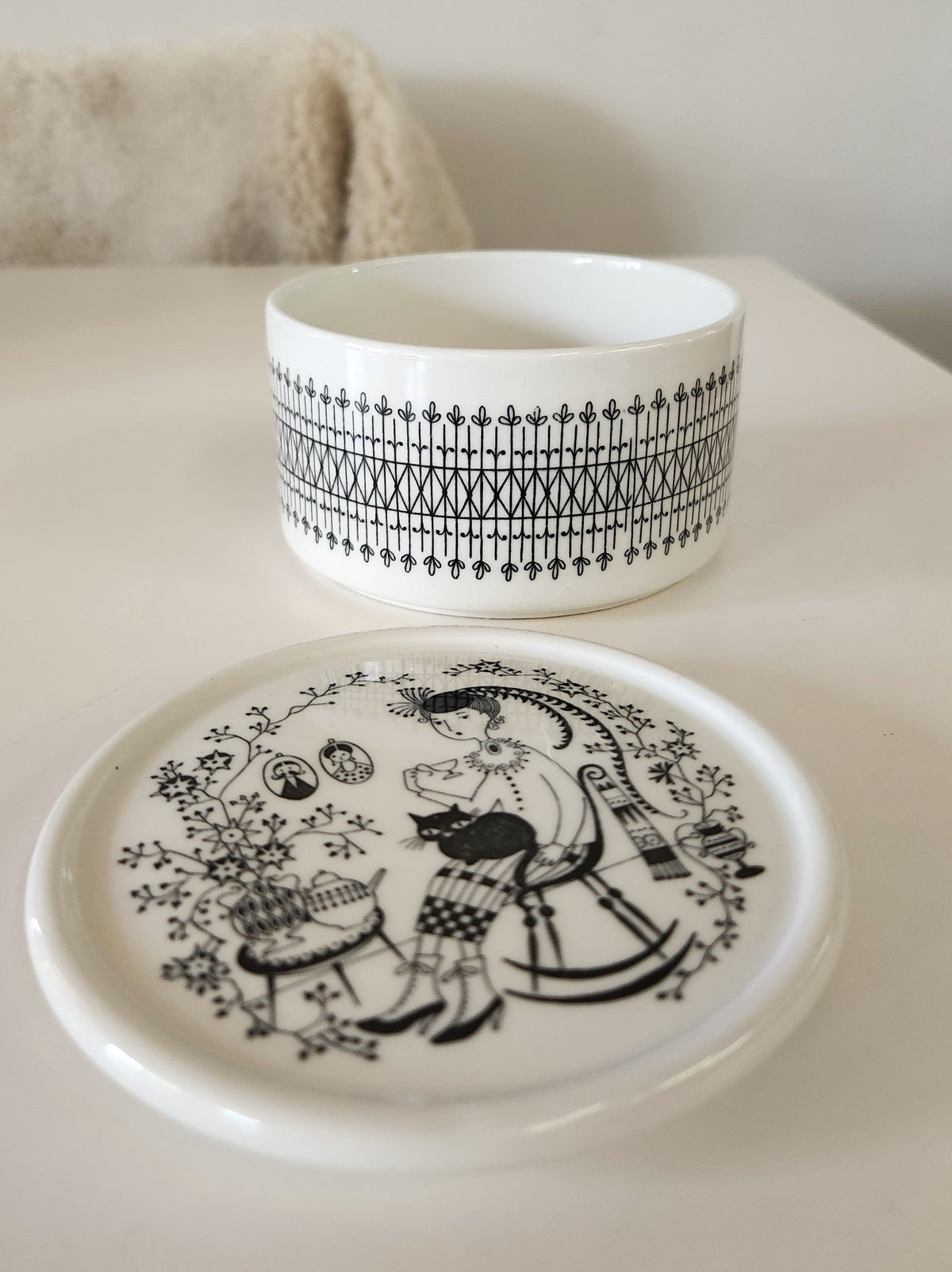 A round white ceramic lidded box made by Arabia, with black hand painted patterns, including a decorative border and a portrait illustration on the lid of a woman with a fat cat on her lap, set against a neutral white background. 