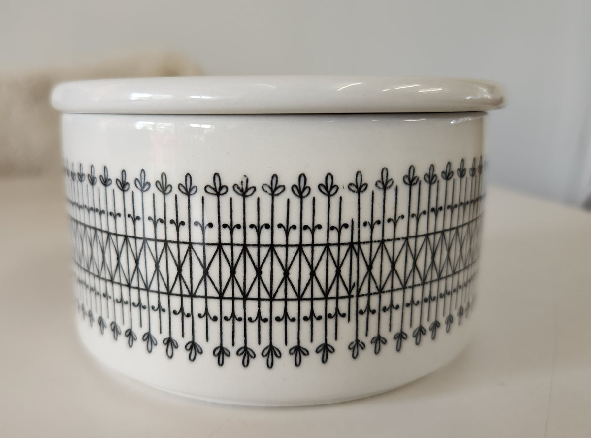 A round white ceramic lidded box made by Arabia, with black hand painted patterns, including a decorative border set against a neutral white background. 