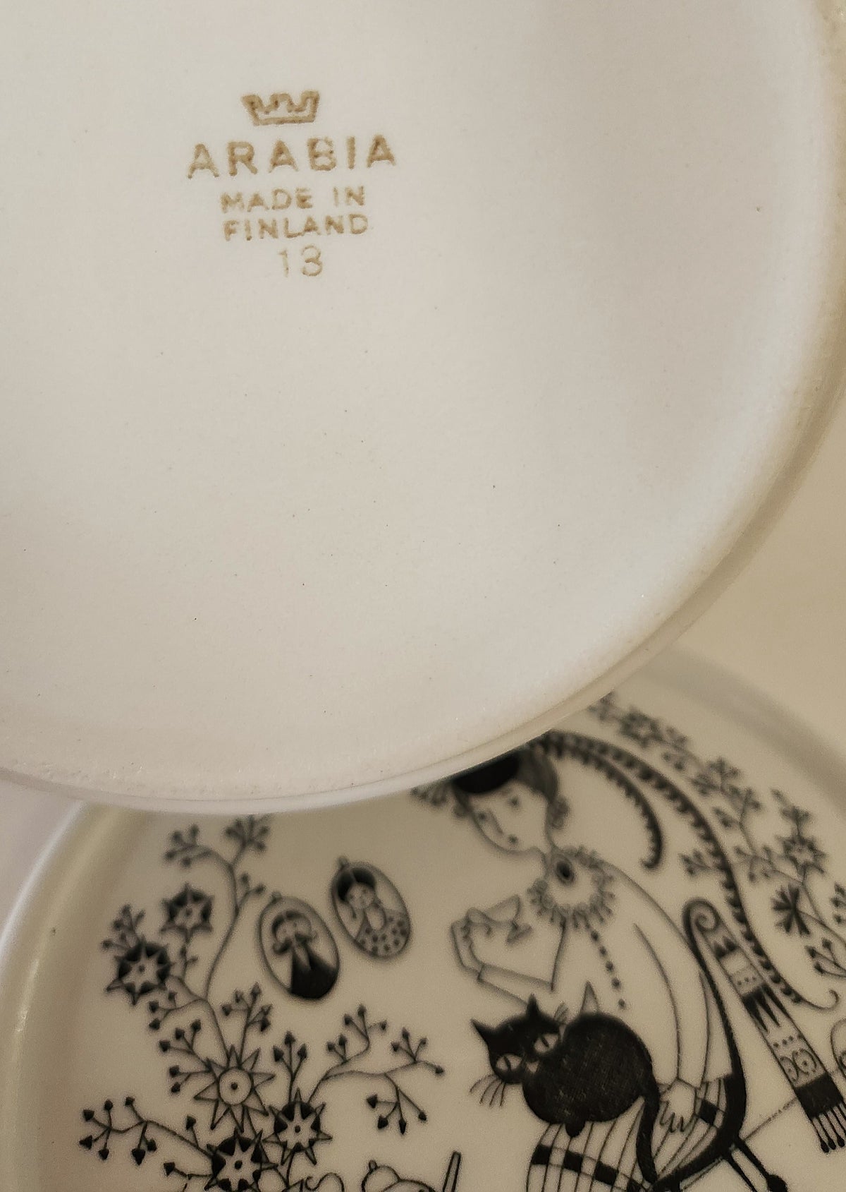 Close-up of the underside of a white ceramic plate or dish with gold 'ARABIA MADE IN FINLAND 13' text and part of a black illustrated floral-and-figure pattern visible on another piece below.