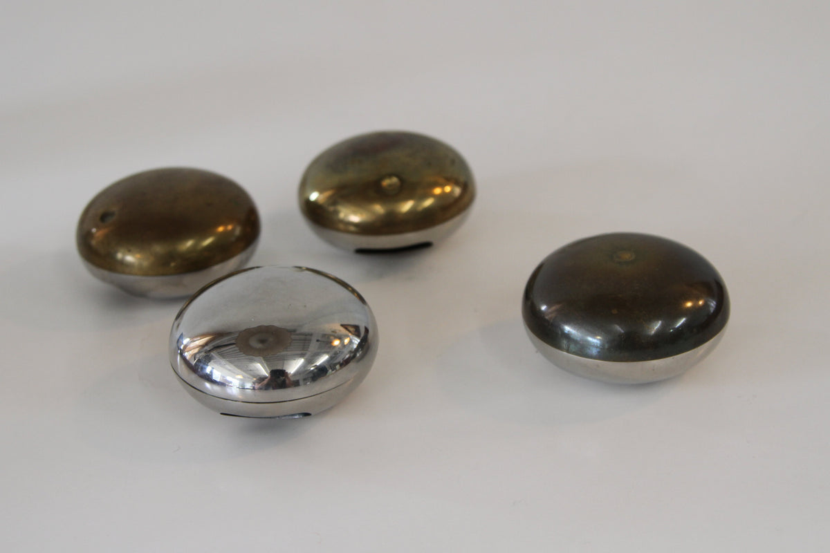 Four small oval compact cases with reflective metallic lids, two in a bronze finish and two in a silver finish, arranged on a plain white surface.