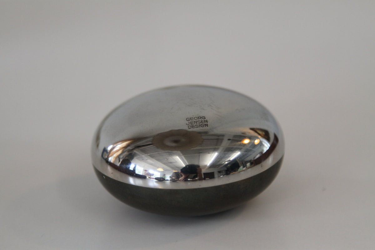 A small, rounded metallic compact case with a reflective silver top and a dark gray base, photographed on a neutral light gray background.