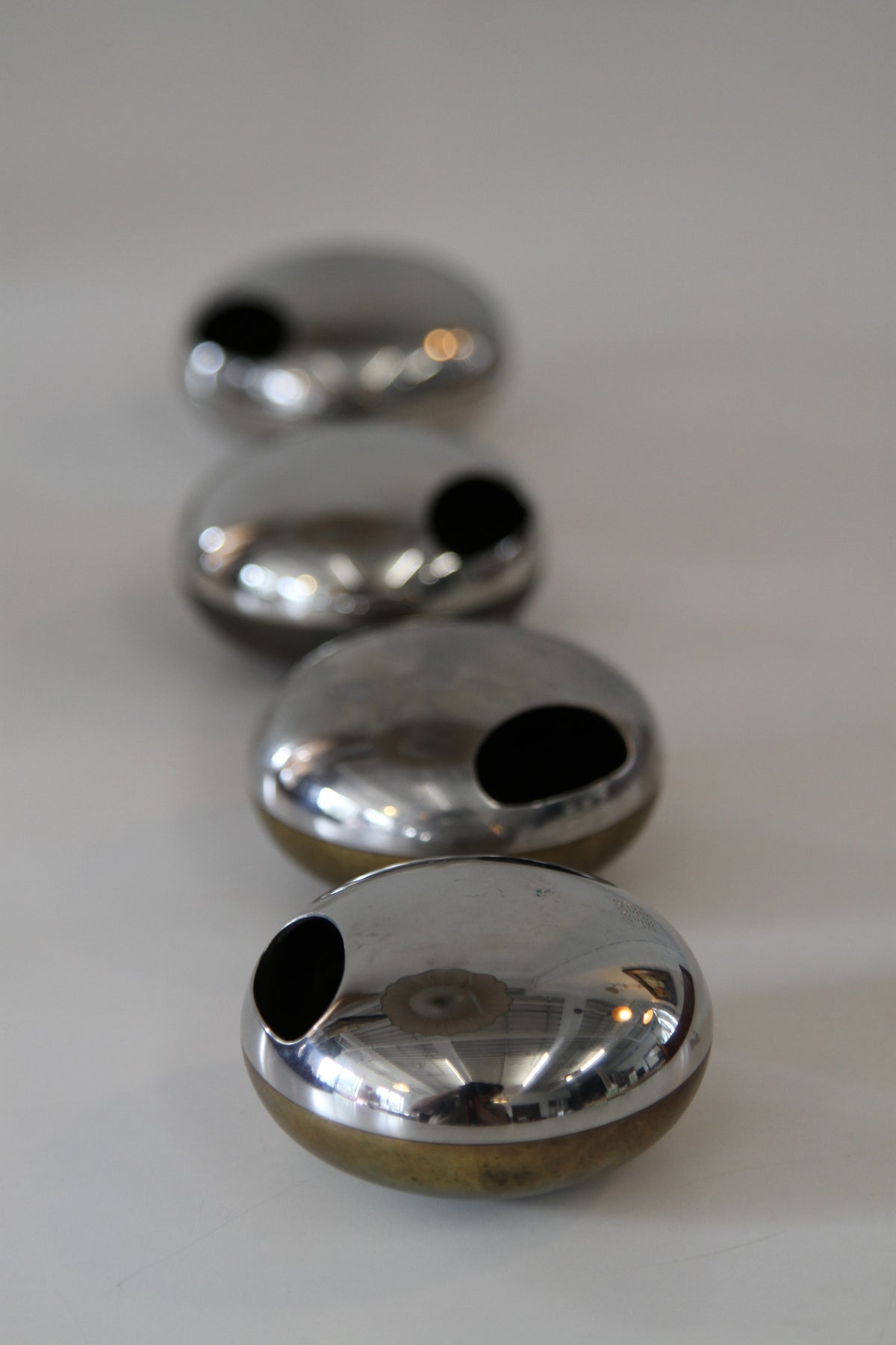 Four small metallic orbs with a glossy reflective silver finish and a black circular cutout on the side, arranged diagonally on a light neutral background.