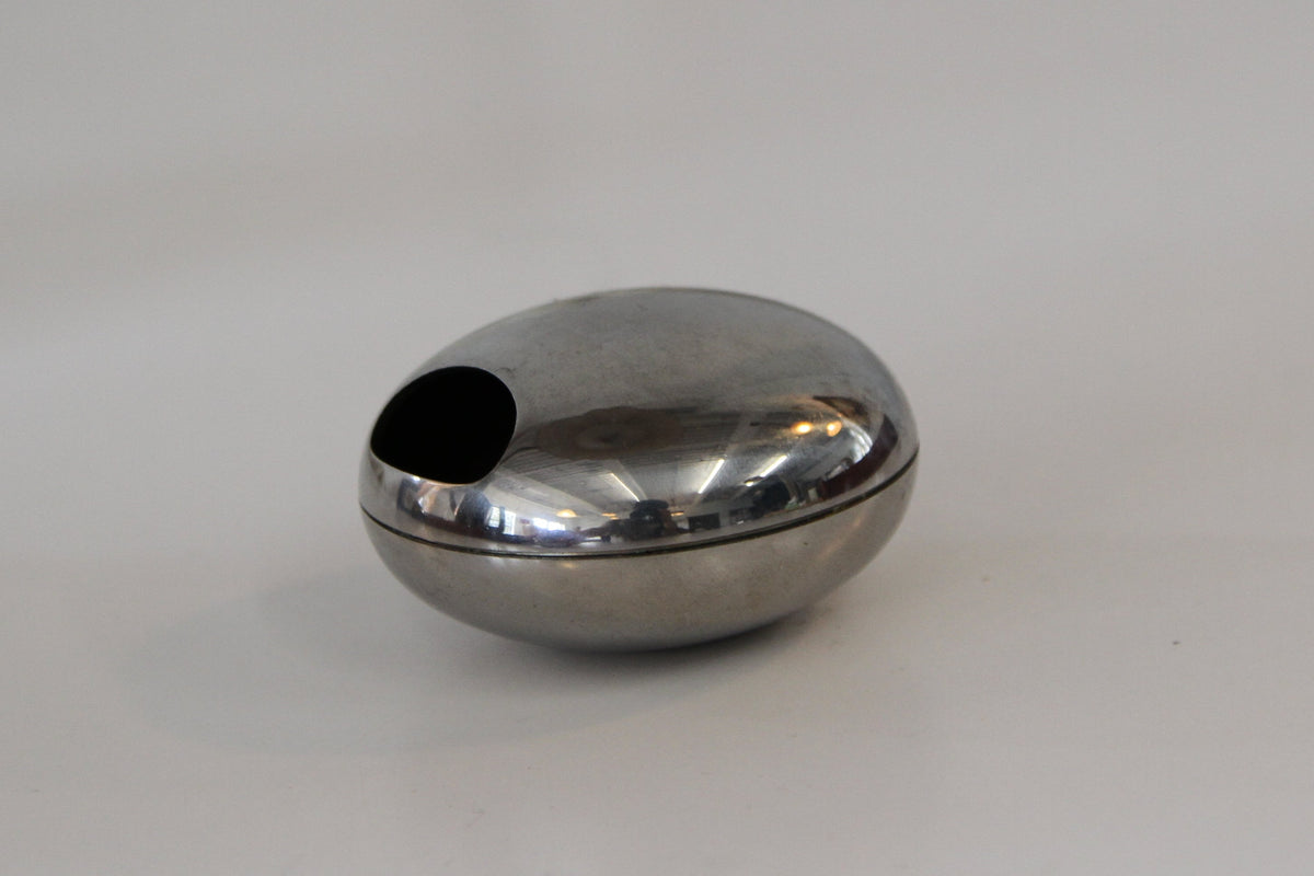 A small polished metallic capsule-shaped object with a dark circular opening on top, resting on a plain white surface.