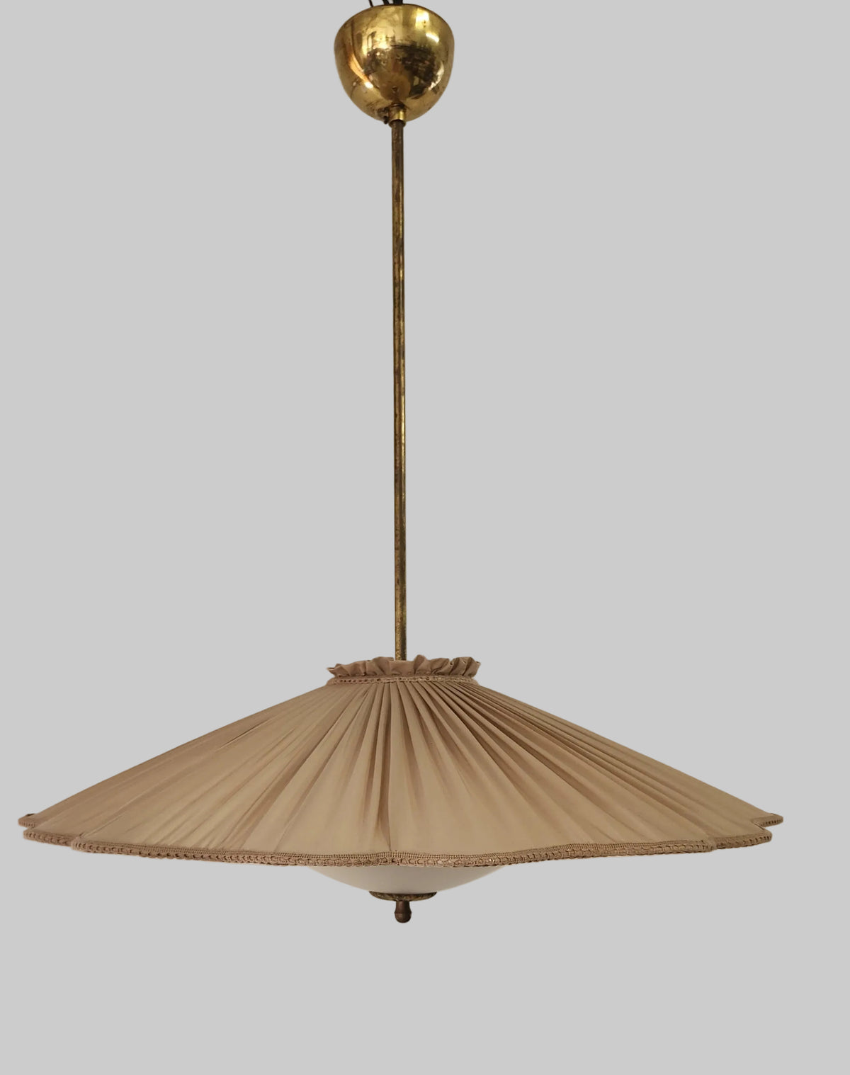 A vintage mid-century modern pendant light with a pleated taupe fabric shade and a brass suspension rod and ceiling canopy, shown hanging against a white wall.