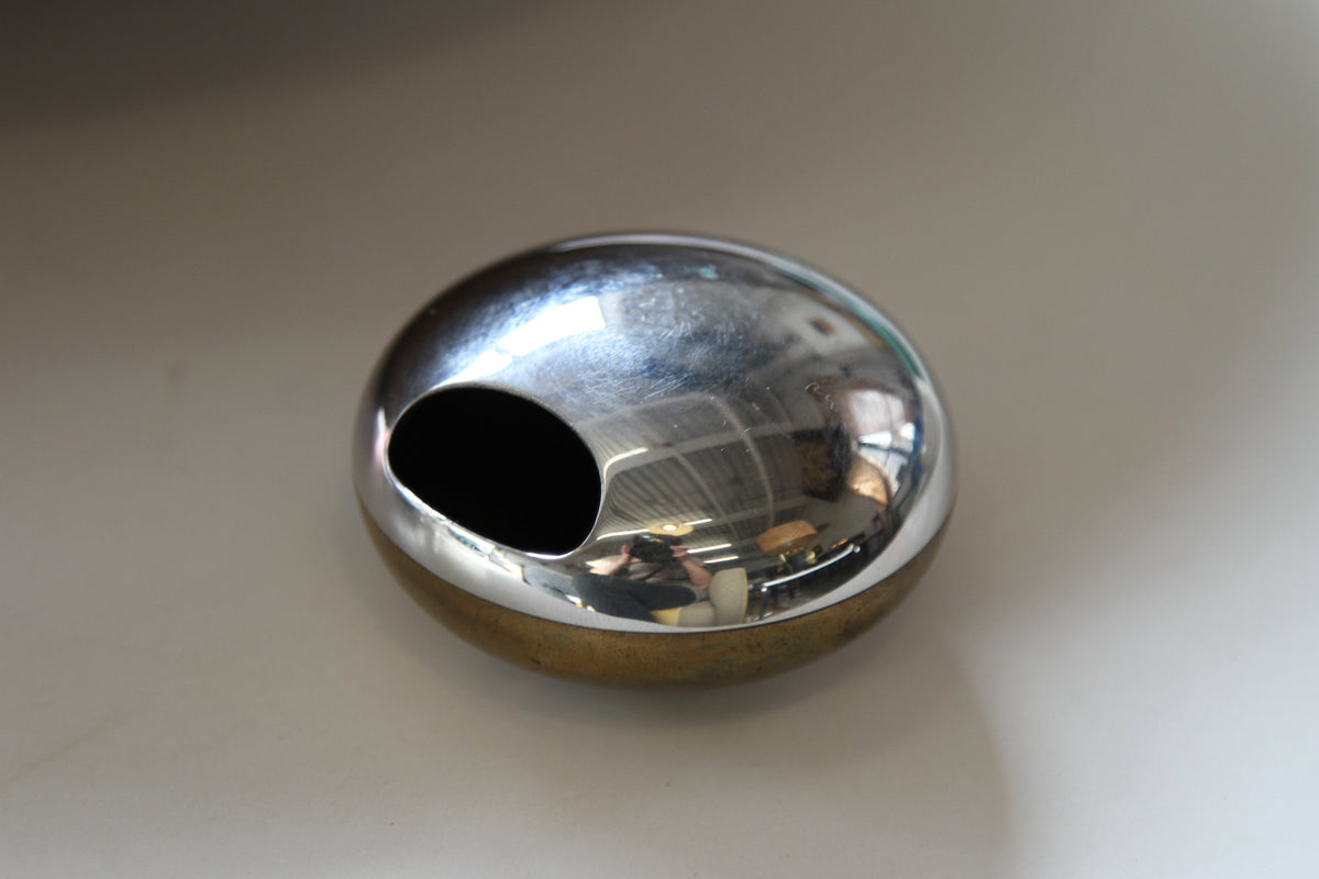 A small polished metallic orb object with a dark oval opening, resting on a plain light surface.