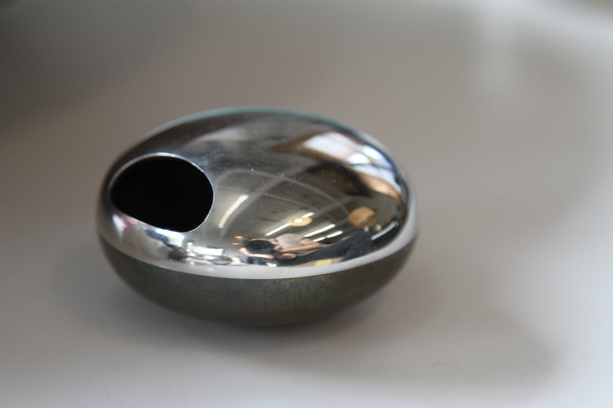 A polished chrome oval soap dish with a circular cutout, resting on a neutral surface. Top is polished silver, base is bronze.