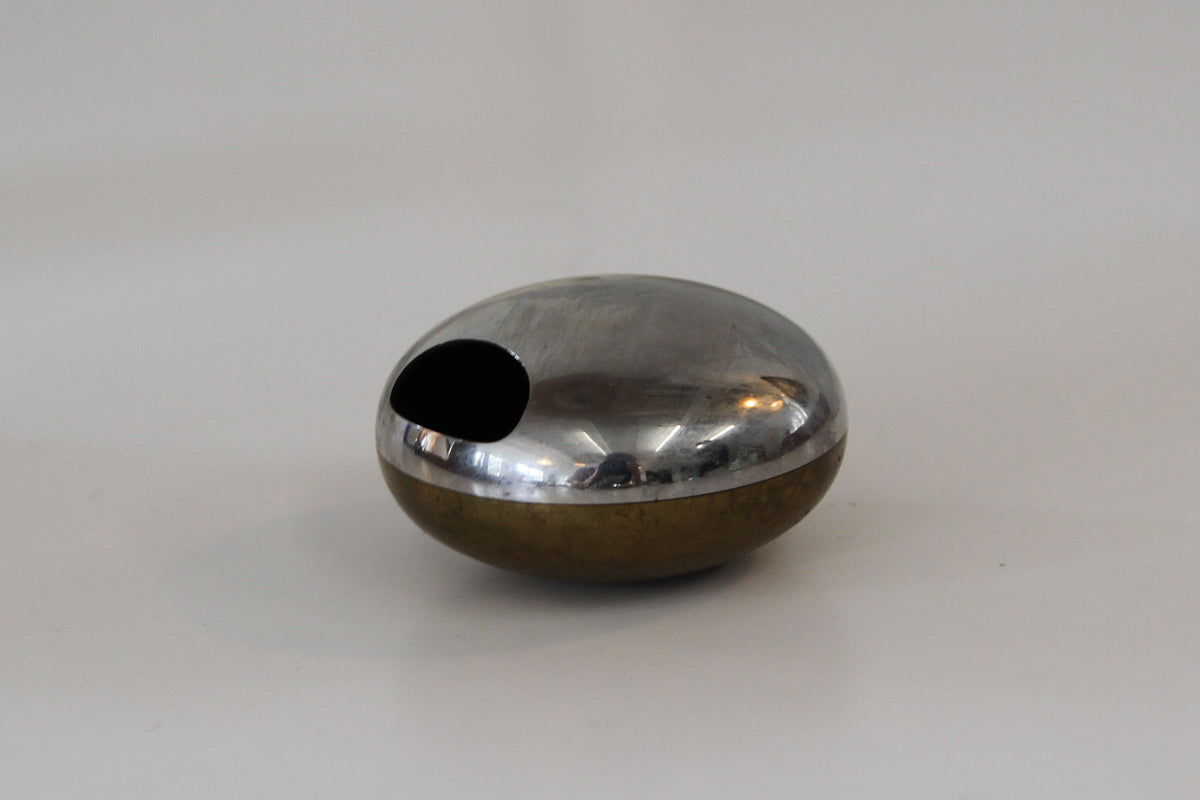 A small metallic capsule-shaped object with a circular cutout, shown on a plain white surface with a soft-focus neutral background.