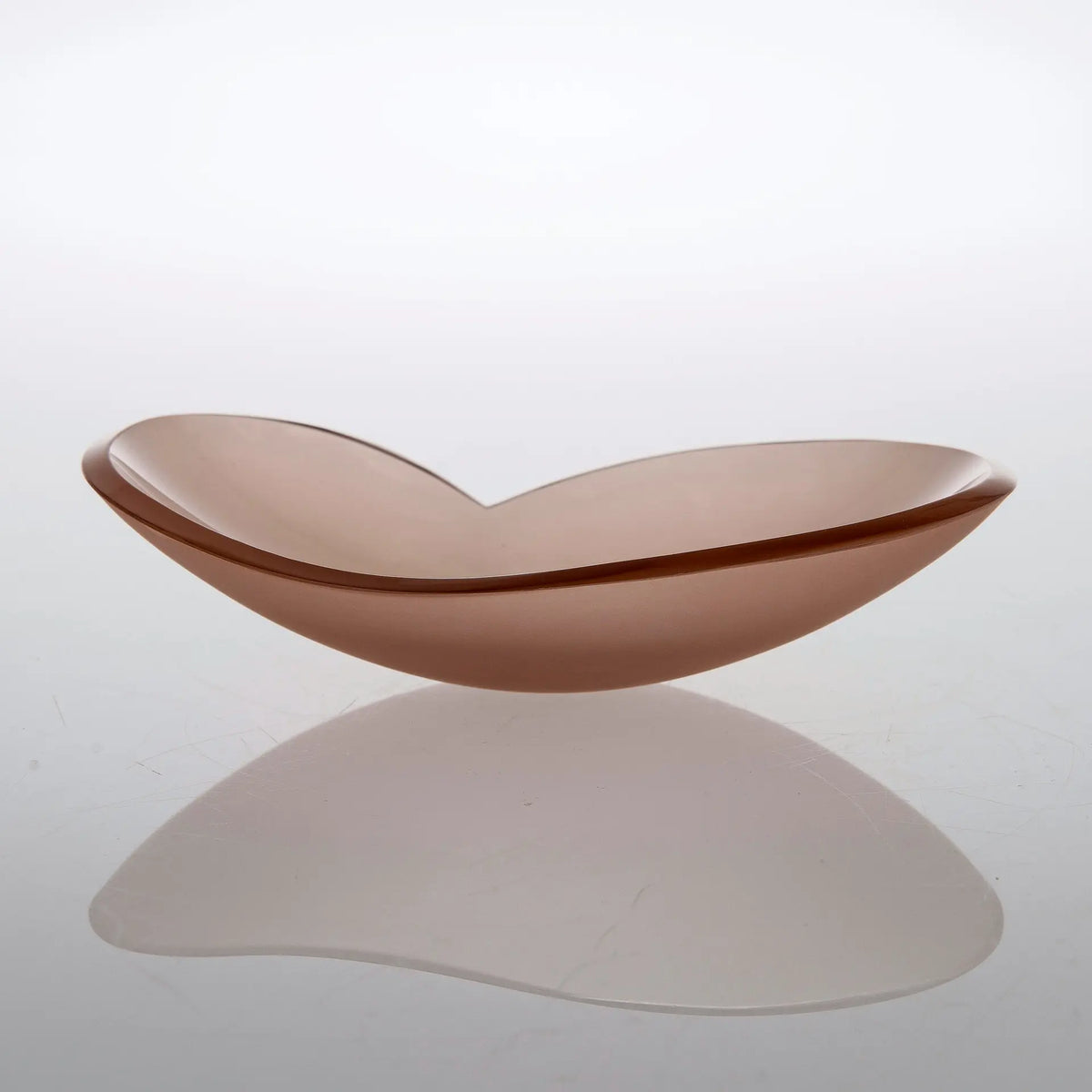 A translucent, organic-shaped glass bowl in a dusty rose or mauve color, resting on a reflective white surface with a soft reflection underneath.