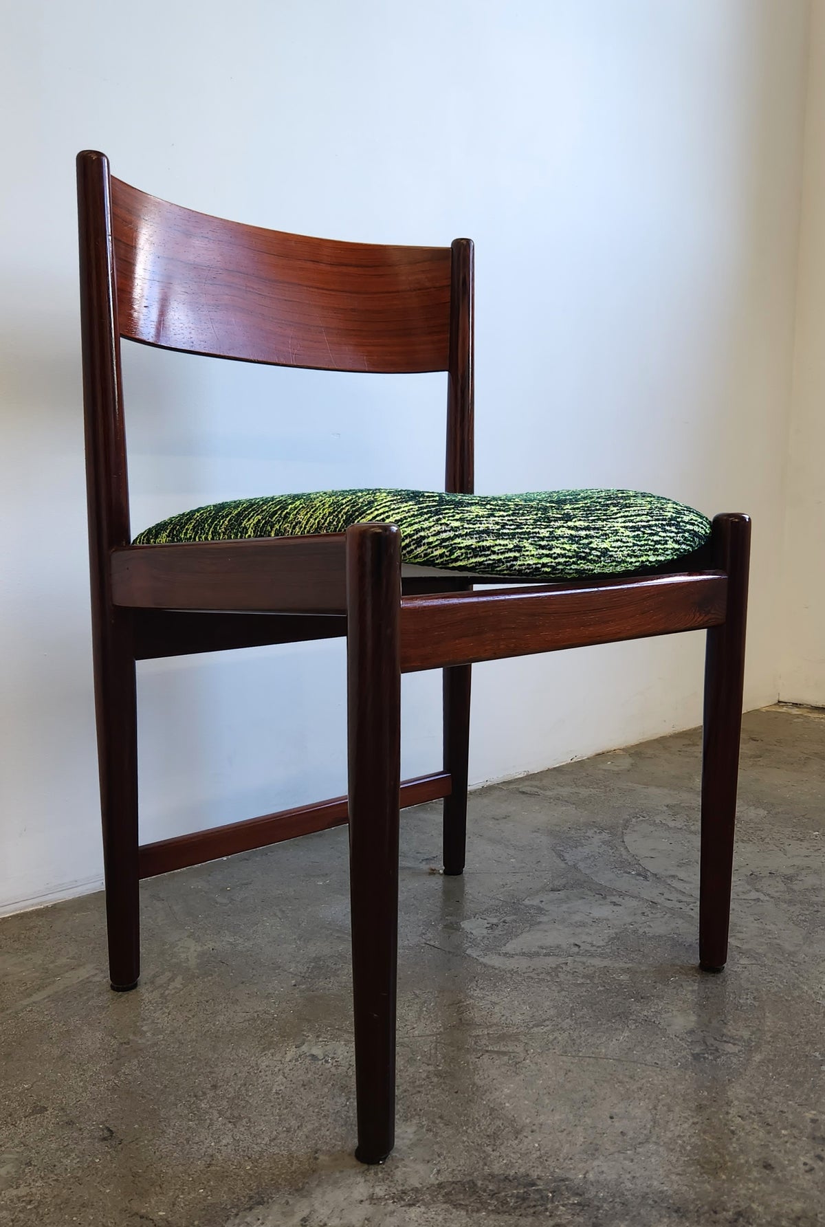 A mid-century modern rosewood wooden dining chair with a dark reddish-brown wood frame and a textured green and black patterned seat cushion, set against a plain off-white wall and concrete floor.