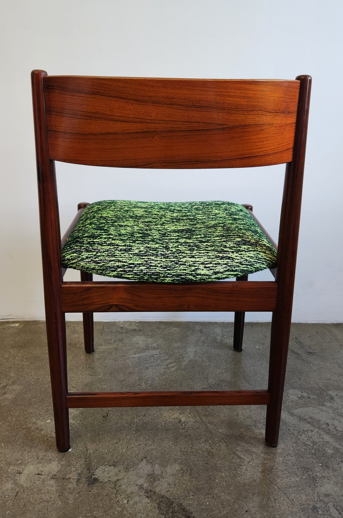 A mid-century modern wooden dining chair with a warm reddish-brown frame and a green marled upholstered seat cushion, set against a plain white wall and concrete floor.