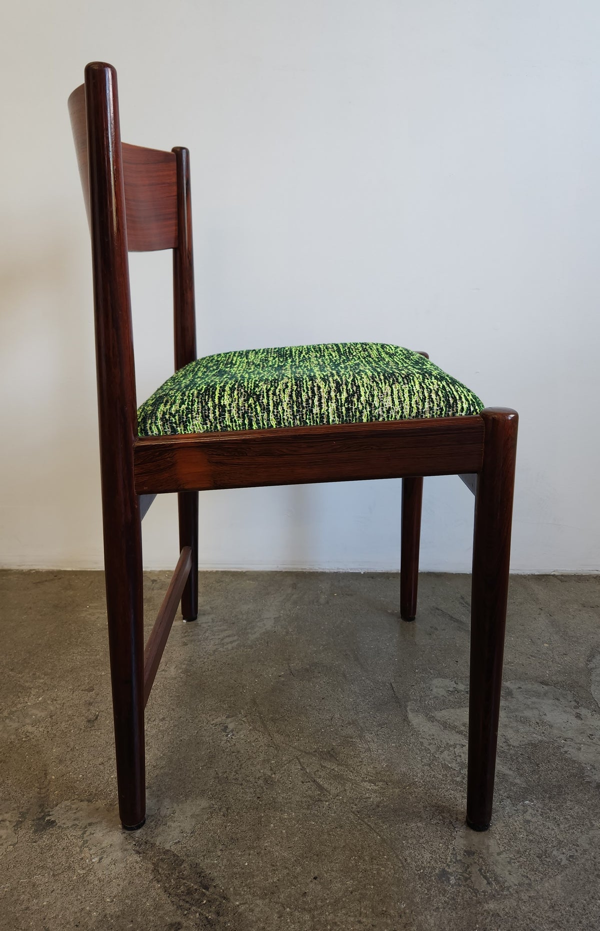A mid-century modern wooden dining chair with a dark reddish-brown frame and a textured green and black patterned seat cushion, shown against a plain white wall and concrete floor.