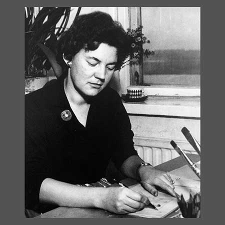 Black and white historical photograph of a Finnish woman seated at a desk, writing or signing a document, with a pin on her dark blouse and paint brushes visible in the foreground.