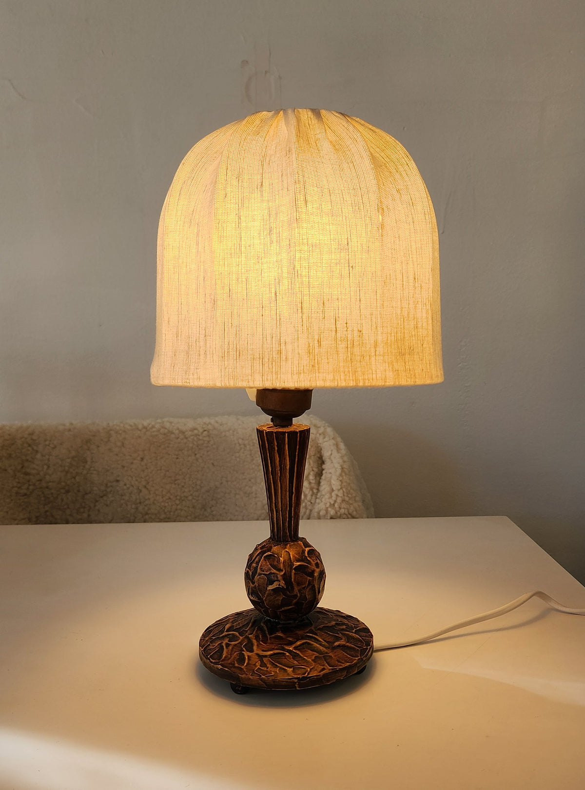 A vintage Swedish wooden table lamp with a warm glowing fabric shade and an ornate dark brown carved base, placed on a white tabletop against a neutral gray wall.