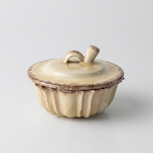 A small lidded ceramic container with a ribbed, pumpkin-like shape and a loop handle on top, shown on a clean light gray studio background.