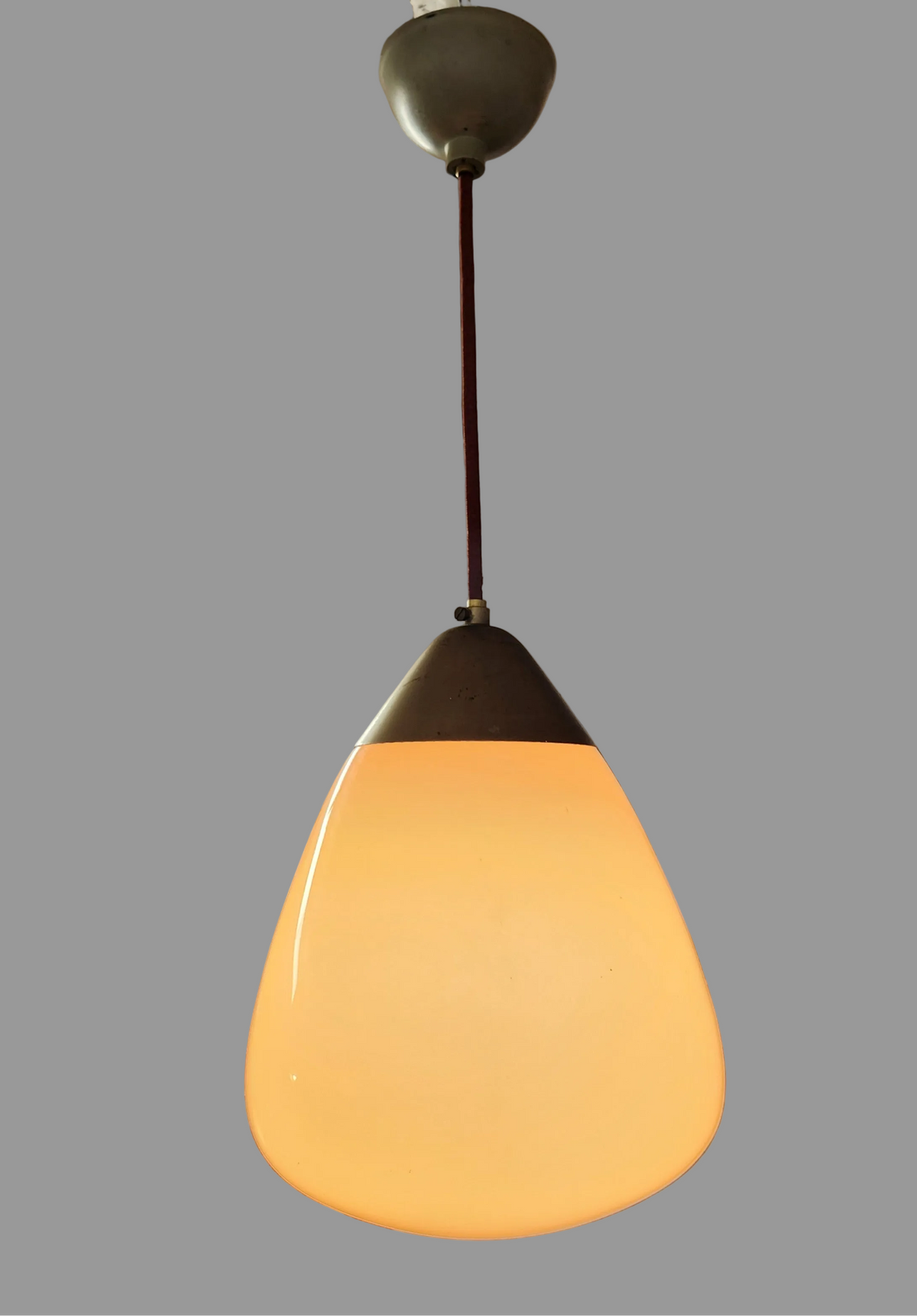 A mid-century modern teardrop pendant light with a glowing warm white glass shade, dark metal collar, and a dark brown hanging cord against a solid gray background.